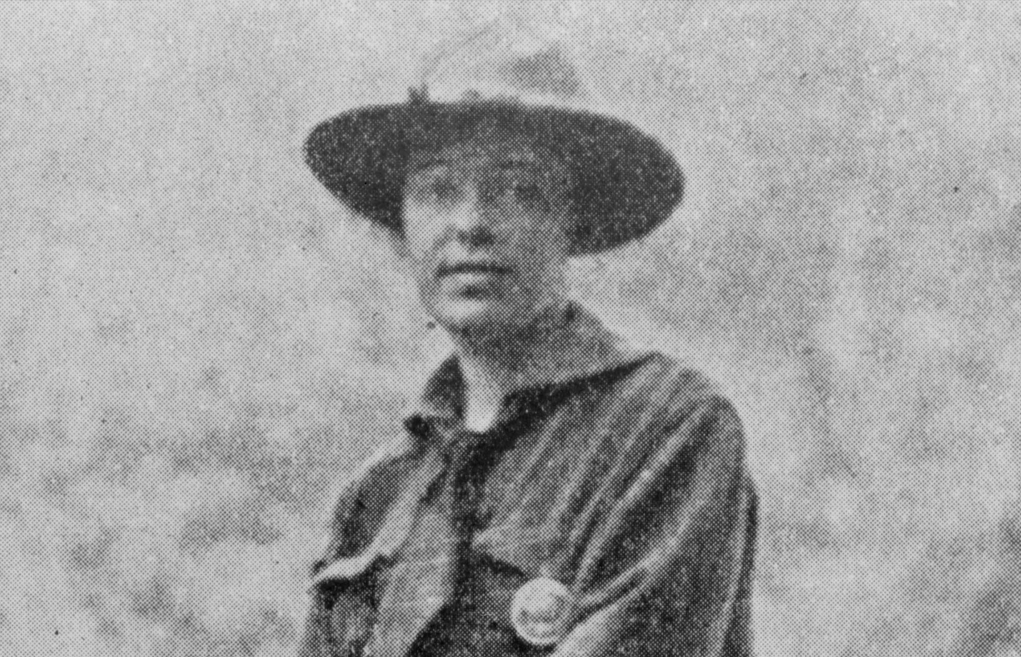 Clare Hodges on horseback wearing a split skirt, shirt, bandana, gloves, and a flat brim hat with a round badge pinned on the pocket.